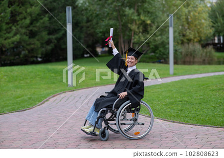Caucasian woman in a wheelchair in a graduate costume rejoices at receiving a diploma. Caucasian woman in a wheelchair in a graduate costume rejoices at receiving a diploma. 102808623