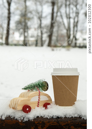 Wooden car carrying Christmas tree with paper cup mock up of coffee or hot chocolate over snow. Copy space for text Toy car in snowy landscape. Merry Christmas and Happy New Year concept 102812300