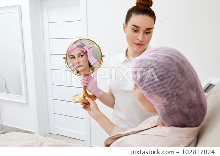 Young woman sitting in medical chair and looking in the mirror. She is satisfied after successful beauty treatment Young woman sitting in medical chair and looking in the mirror. She is satisfied after successful beauty treatment 102817024
