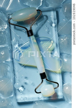 Blue jade face roller and Gua Sha scraping for beauty facial massage therapy on ice cubes and water background. 102819046