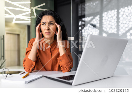 Beautiful woman at workplace overworked, tired hispanic woman at workplace with severe headache, businesswoman working inside office sitting with laptop, stressed and sad. 102820125