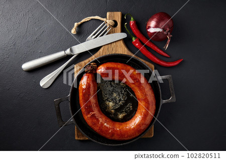 Fried sausage rolled into a ring on a cast iron pan with cutlery, a wooden cutting board, hot peppers and red onions 102820511