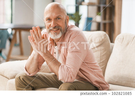Smiling, happy senior man sitting on a sofa in the comfort of his home. With his hands held together, he radiates a sense of contentment and relaxation. . High quality photo Smiling, happy senior man sitting on a sofa in the comfort of his home. With his hands held together, he radiates a sense of contentment and relaxation. . High quality photo 102821085