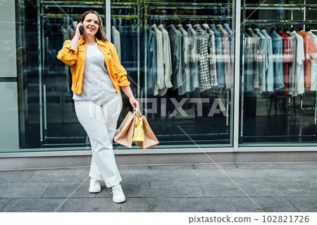 Plus size woman with shopping bags talking by cell phone on shop window background. Shopping woman talking on the smartphone 102821726