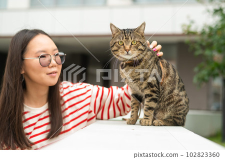 A striped cat sits on a bench outdoors. Young woman walking with her pet outdoors. 102823360