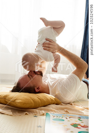 Happy dad holds his newborn baby in his arms while lying on the floor in a bright child's room. The father looks at the child with a loving look. Happy childhood and fatherhood. Happy dad holds his newborn baby in his arms while lying on the floor in a bright child's room. The father looks at the child with a loving look. Happy childhood and fatherhood. 102824596