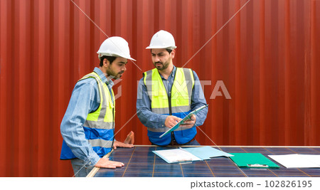 Shipment worker pointing at shipping schedule on clipboard, explain to colleague about planning for next shipment. They use solar cell panel as a table. A large container is in the background Shipment worker pointing at shipping schedule on clipboard, explain to colleague about planning for next shipment. They use solar cell panel as a table. A large container is in the background 102826195