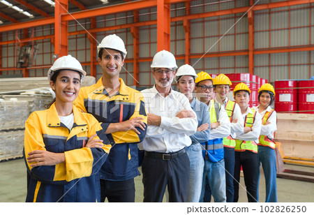 Group of male and female factory labor stand smiling together with arms crossed in industry factory. Everyone wearing safety uniform and helmet. Workers working in the metal sheet factory. 102826250