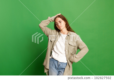 Portrait with one doubtful young girl, teenager in casual clothes touching head and looking away with pensive face over green color studio background 102827502