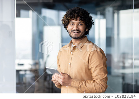Portrait of young programmer with tablet computer inside office, hispanic man with curly hair smiling and looking at camera, man at workplace testing online applications standing near window Portrait of young programmer with tablet computer inside office, hispanic man with curly hair smiling and looking at camera, man at workplace testing online applications standing near window 102828194