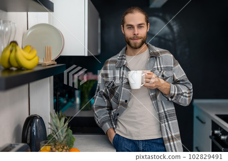 Man with beard holding cup with coffee, standing by kitchen counter with modern light interior. Man with beard holding cup with coffee, standing by kitchen counter with modern light interior. 102829491