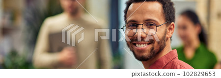 Young cheerful mixed race man holding digital tablet and smiling at camera while standing in the modern office with his colleagues on the background Young cheerful mixed race man holding digital tablet and smiling at camera while standing in the modern office with his colleagues on the background 102829653