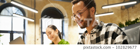 Young smart guy wearing eyeglasses working on laptop while sitting at desk with his female colleague in the modern coworking space or office 102829677