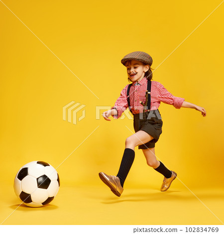 Portrait of stylish little girl, child in retro clothes, cap, shirt and shorts playing with football ball against yellow studio background 102834769