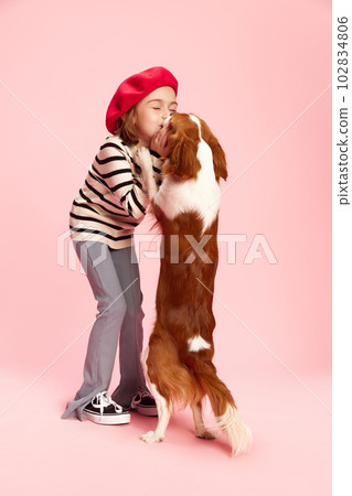 Full-length portrait of tender, caring, little girl, child in red beret and striped sweater playing, kissing dog against pink studio background 102834806