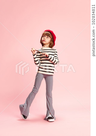 Full-length portrait of dreamy little girl, child in red beret and striped sweater posing with painting brush against pink studio background 102834811