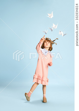 Full-length portrait of playful, lovely little girl in pink dress posing, playing with paper birds against blue studio background Full-length portrait of playful, lovely little girl in pink dress posing, playing with paper birds against blue studio background 102834814