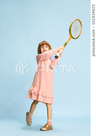 Full-length portrait of little girl in pink dress posing with tennis racket, playing against blue studio background 102834823