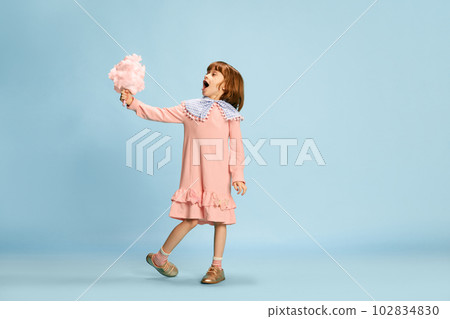 Portrait of happy little girl, child in cute pink dress holding candy floss, standing against blue studio background. Cheerful time 102834830