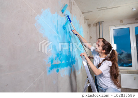 Couple in new home during repair works painting wall together. Happy family holding paint roller painting wall with blue color paint in new house. Home renovation DIY renew home concept Couple in new home during repair works painting wall together. Happy family holding paint roller painting wall with blue color paint in new house. Home renovation DIY renew home concept 102835599