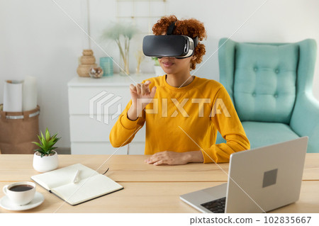 African american young woman wearing using virtual reality metaverse VR glasses headset at home. Girl touching air during VR experience on virtual reality helmet. Simulation hi-tech videogame concept 102835667