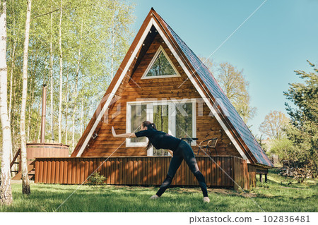 Woman does stretching on lawn in front of her a-frame Woman does stretching on lawn in front of her a-frame 102836481