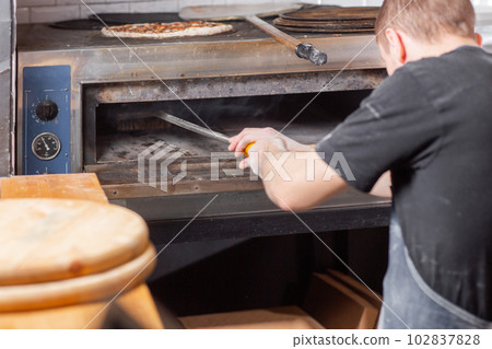 The chef cleaning the pizza oven. Raw pizza ready to bake. Cook in a blue apron in the kitchen. with a shovel in his hands. boxes for food delivery on background. The chef cleaning the pizza oven. Raw pizza ready to bake. Cook in a blue apron in the kitchen. with a shovel in his hands. boxes for food delivery on background. 102837828