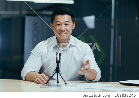 Asian smiling young man sitting in office at desk in front of camera, talking on microphone via video call, blogging, webinar, training, recording podcast. 102838249