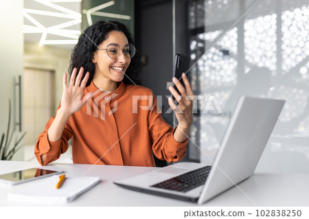 Young Muslim woman sitting in the office at the table, working on the laptop and talking on the phone on a video call, smiling, greeting, waving at the camera. Young Muslim woman sitting in the office at the table, working on the laptop and talking on the phone on a video call, smiling, greeting, waving at the camera. 102838250