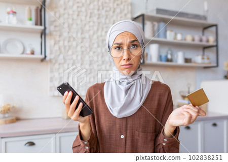 Upset young muslim woman wearing hijab feeling frustration and problems with credit card account and finances. He spreads his hands, holds the phone, looks worriedly into the camera. Upset young muslim woman wearing hijab feeling frustration and problems with credit card account and finances. He spreads his hands, holds the phone, looks worriedly into the camera. 102838251