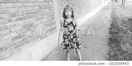Girl scared. Halloween theme. A child in a dress with flowers against a brick wall is afraid. The girl sees a zombie on the street, is surprised and she is afraid. Black and white monochrome image 102838443