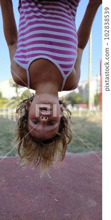 Girl upside down. A child with blond hair in a striped pink dress hangs from a bar in the playground. The hair hangs down, the mouth is half open. Girl looking at the camera 102838539