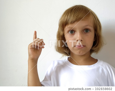 A little cute girl 6-7 years old with blond hair on a white background, pointing up. A child with tanned skin. White T-shirt. Copy space. Girl with her hand points to something. Bob hairstyle A little cute girl 6-7 years old with blond hair on a white background, pointing up. A child with tanned skin. White T-shirt. Copy space. Girl with her hand points to something. Bob hairstyle 102838602
