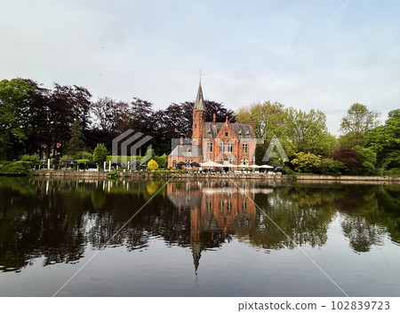 Minnewater ancient medieval gothic red brick castle at the Lake of Love in Bruges in public park zone, Belgium, bright sunny day, beautiful natural landscape, building reflection in water 102839723