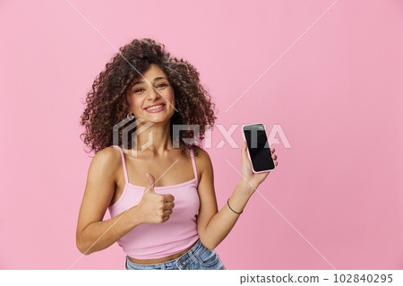 Woman blogger holding phone video call takes selfies, with curly hair in pink smile t-shirt and jeans poses on pink background, copy space, technology and social media, online 102840295