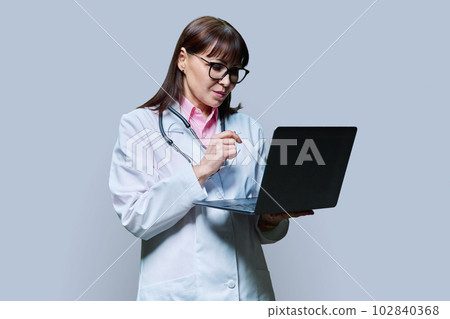 Mature female doctor using laptop on gray background Mature female doctor using laptop on gray background 102840368