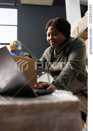 Stockroom worker analyzing online orders on laptop computer, preparing clients packages for shipping using cardboard boxes in storage room. African american employee working in warehouse 102841381