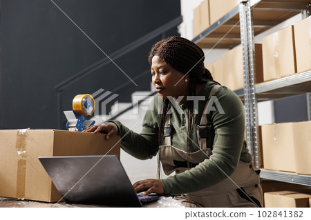 Stockroom supervisor preparing customers orders using cardboard boxes, checking shipping details on laptop computer in storehouse. African american employee working at products inventory report Stockroom supervisor preparing customers orders using cardboard boxes, checking shipping details on laptop computer in storehouse. African american employee working at products inventory report 102841382