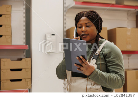 Storehouse employee talking at landline phone with remote manager, discussing products inventory in storage room. Supervisor wearing industrial overall checking online orders on tablet computer 102841397