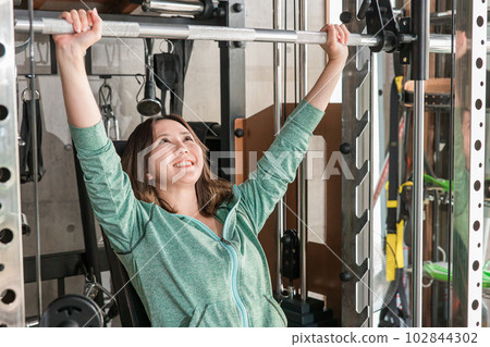 A smiling Japanese woman doing muscle training using a shoulder press machine at the gym A smiling Japanese woman doing muscle training using a shoulder press machine at the gym 102844302