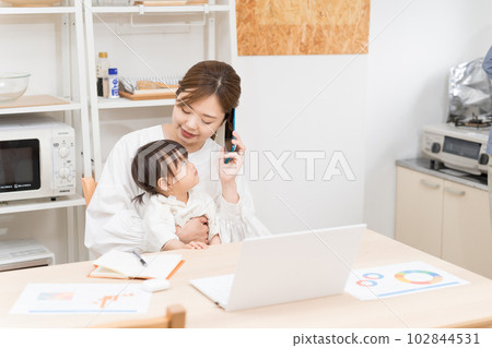 一位年輕女性在餐廳裡抱著嬰兒,一邊打電話一邊在家工作 一位年輕女性在餐廳裡抱著嬰兒,一邊打電話一邊在家工作 102844531