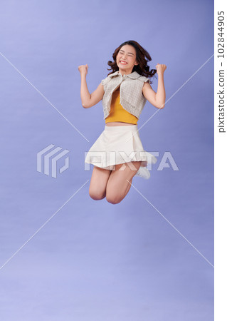 Full length of cheerful young woman jumping high smiling showing power muscles isolated on purple background Full length of cheerful young woman jumping high smiling showing power muscles isolated on purple background 102844905