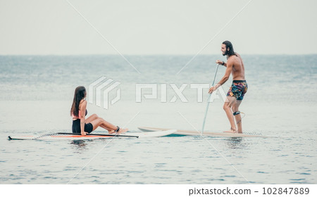 Sea woman and man on sup. Silhouette of happy young woman and man, surfing on SUP board, confident paddling through water surface. Idyllic sunset. Active lifestyle at sea or river. 102847889