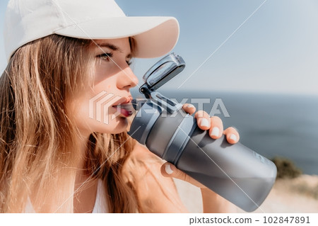Fintess woman drinking water. Happy, active middle aged woman standing on beach and drinking water after excersise. Concept of lifestyle, sport. Close up Fintess woman drinking water. Happy, active middle aged woman standing on beach and drinking water after excersise. Concept of lifestyle, sport. Close up 102847891