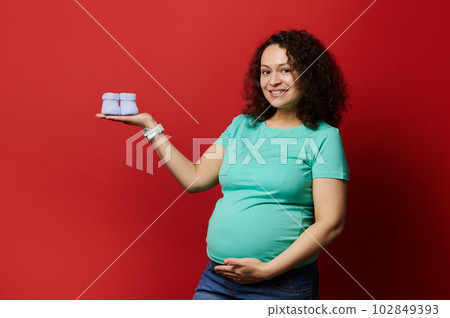 Expectant pregnant mother, gravid woman holding blue knitted baby booties socks for hew newborn baby boy in comb, isolated red background. Copy space. Newborn clothing. Pregnancy fashion. Maternity 102849393