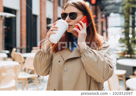 Happy cheerful young redhead woman in fashion office clothes and sunglasess talking on Phone with eco cup of tea. Mockup, Woman holding smartphone and bio coffee cup. Urban concept 102852097