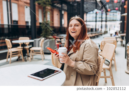 Young woman enjoying a coffee, sitting on the cafe terrace on the modern city street. Person sitting at table and using smartphone outdoors. Online education, order, working or shopping concept 102852112