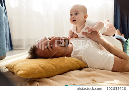 Happy dad holds his newborn baby in his arms while lying on the floor in a bright child's room. The father looks at the child with a loving look. Happy childhood and fatherhood. Happy dad holds his newborn baby in his arms while lying on the floor in a bright child's room. The father looks at the child with a loving look. Happy childhood and fatherhood. 102853628