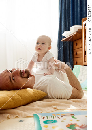 Happy dad holds his newborn baby in his arms while lying on the floor in a bright child's room. The father looks at the child with a loving look. Happy childhood and fatherhood. 102853631