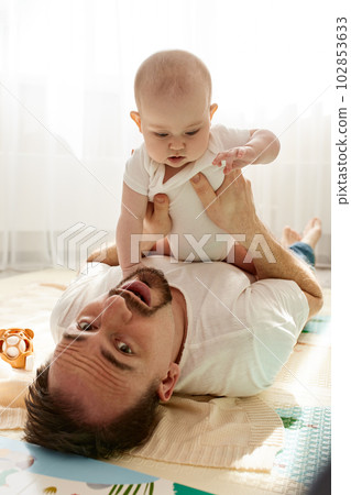 Happy dad holds his newborn baby in his arms while lying on the floor in a bright child's room. The father looks at the child with a loving look. Happy childhood and fatherhood. Happy dad holds his newborn baby in his arms while lying on the floor in a bright child's room. The father looks at the child with a loving look. Happy childhood and fatherhood. 102853633